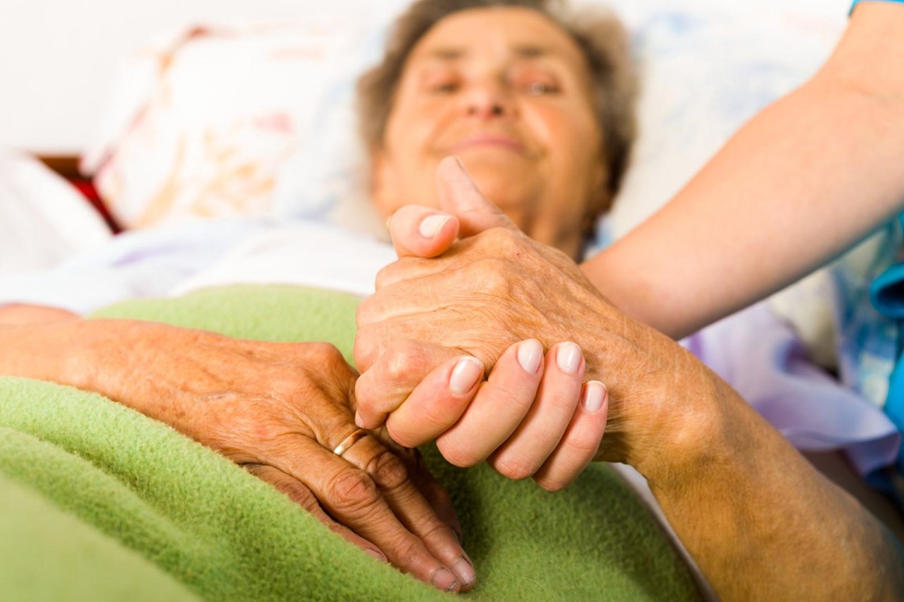 jüngere Hand hält ältere Hand, im Hintergrund das Gesicht einer alten Frau